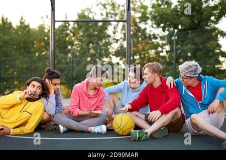 relaxed male teens have rest after sport games, basketball outdoors in court. caucasian boys in casual wear enjoy spending time with friends Stock Photo