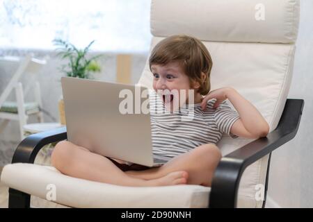 Happy surprised kid boy sit on computer using laptop and having a video call. Smiling child sitting at home rest on chair busy with gadgets. Online Stock Photo