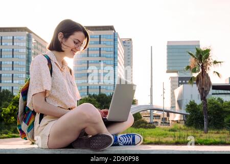 young traveler woman with backpack sitting in the city typing on her computer, concept of technology, youth and digital nomad lifestyle Stock Photo