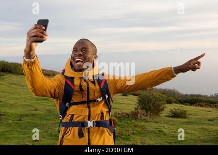Portrait of happy african american hiker taking selfie and pointing at view Stock Photo