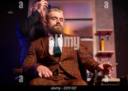 young caucasian barber cutting hair of his cool rich client sitting in armchair and smoking cigar, man sitting in barberchair, he likes getting haircut. barbershop concept Stock Photo
