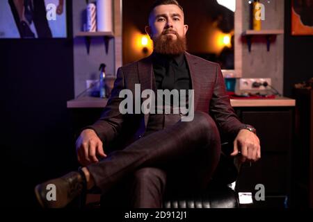 self-confident brutal male in tuxedo is in contemplation indoors, elegant male sits on chair posing Stock Photo