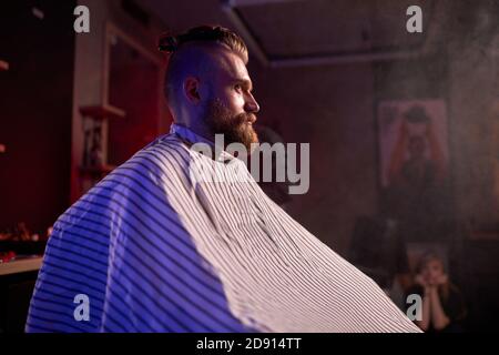 side view on young bearded male sitting in barber shop, waiting for barber, looking away, in apron Stock Photo