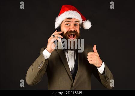 Christmas celebration concept. Businessman with cheerful face holds mobile phone. Man in smart suit and Santa hat on brown background. Manager with beard talks on cell phone showing thumbs up Stock Photo
