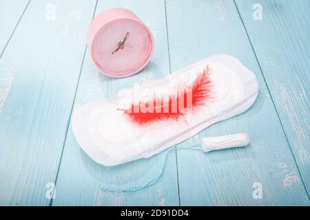 Red feather on a sanitary pad, a tampon and an alarm clock on a blue wooden table Stock Photo