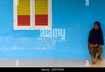 Woman sitting against blue wall with yellow/red shutters,Ternate, North-Moluccas, Indonesia. Stock Photo