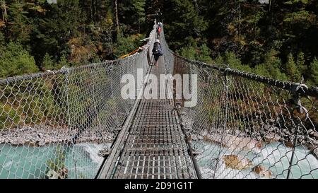 Porters crossing a suspension bridge with attached Buddhist prayer flags above Dudhkoshi River in a valley near Manjo on Mount Everest Base Camp Trek. Stock Photo