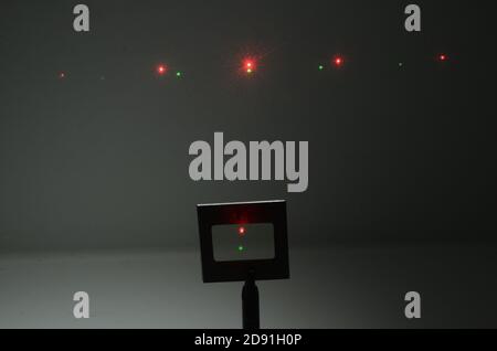 In a darkened room, two laser beams from a source under the camera, hits an optical grid and an interference pattern apppears on the wall in the backg Stock Photo