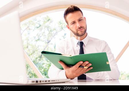 Thinking businessman holding binder in his hand and using laptop while sitting at desk in the office. Stock Photo