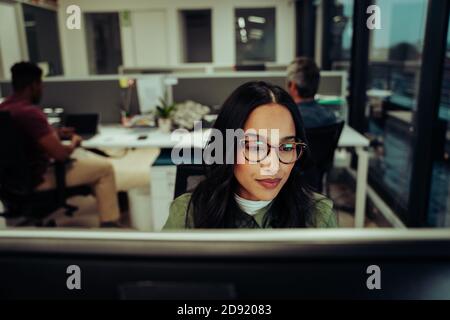 Multi-ethnic female entrepreneur wearing spectacles working on business report typing on desktop computer sitting in office  Stock Photo