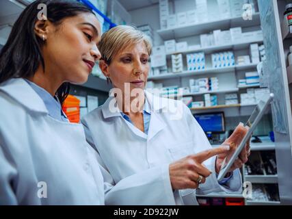 Elderly pharmacist guiding new female intern teaching scrolling on digital tablet standing in pharmacy  Stock Photo