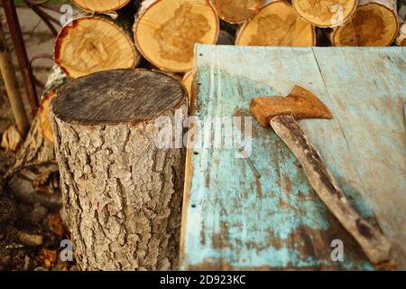 Old rusty ax lies on the blue shabby table near logs of birch wood. Stock Photo