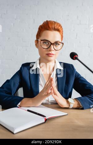 Focused redhead woman fingertips touching gesture, looking away, while sitting at table in boardroom Stock Photo