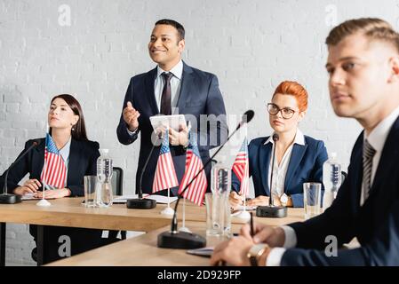 Happy indian politician looking away, while standing with digital tablet near colleagues in boardroom Stock Photo