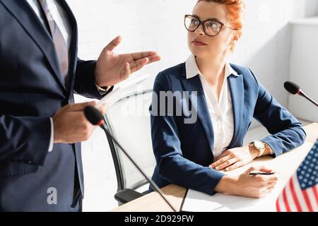 Female politician in eyeglasses looking at colleague, while sitting with pen and clipboard during political party meeting Stock Photo