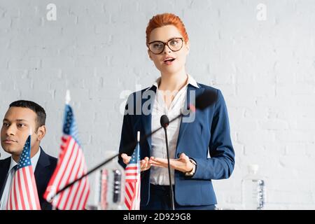 Redhead female politician speaking on political party congress, standing near indian colleague on blurred foreground Stock Photo