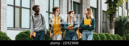 Panoramic shot of smiling multicultural teenage friends with notebooks walking on urban street Stock Photo