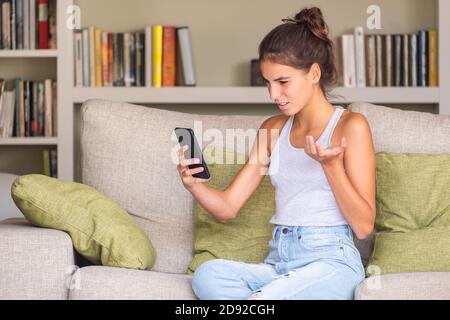 upset young girl reading a message in a smart phone sitting in a sofa at home Stock Photo