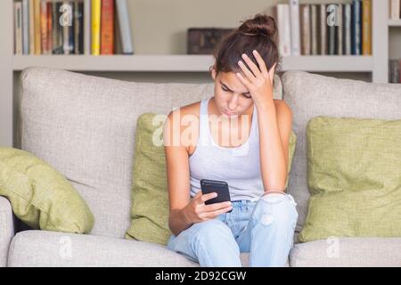 upset young girl reading a message in a smart phone sitting in a sofa at home Stock Photo