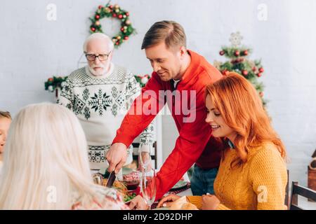 Smiling man cutting turkey on festive table with dinner near family at home Stock Photo