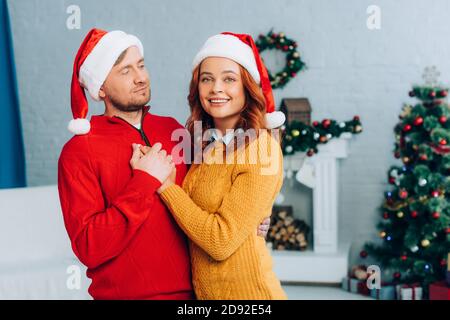 happy woman in santa hat looking at camera while embracing husband on christmas Stock Photo