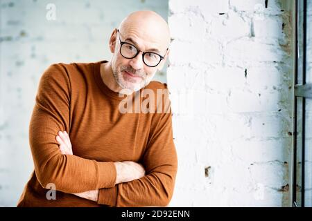 Portrait shot of curious man with beard and eyewear standing at the wall and looking at camera. Stock Photo