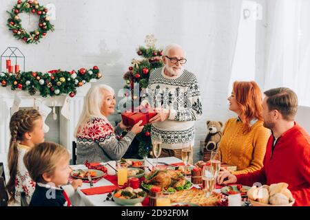 Happy senior couple holding gift box near family sitting at festive table Stock Photo