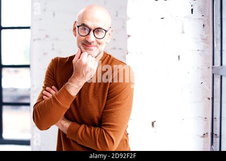 Portrait of curious man with beard and eyewear standing at the window. Hand on chin. Stock Photo