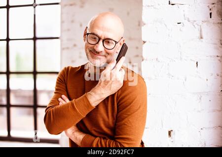 Portrait shot of happy senior man wearing casual clothes while standing by the window and talking with somebody on his mobile phone. Stock Photo
