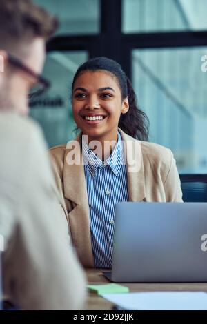Vertical shot of a young cheerful mixed race female office worker working on laptop in the modern office, talking with colleague and smiling Stock Photo