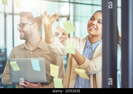 Project planning. Two creative diverse coworkers putting colorful sticky notes on a glass wall while standing in the modern office Stock Photo