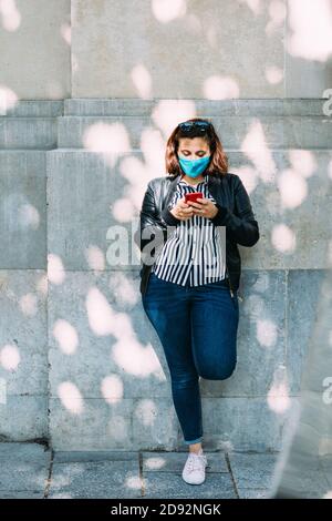 woman with a face mask leaning against the wall using her smartphone Stock Photo