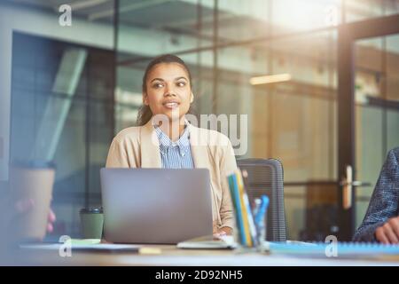 Young mixed race female office worker working on laptop in the modern office, looking aside and thinking about something Stock Photo