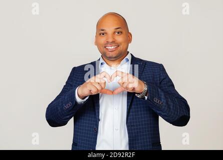 Portrait of a romantic young African American businessman guy smiling in love showing heart symbol and shape with hands. Romantic concept. Standing on a gray background Stock Photo