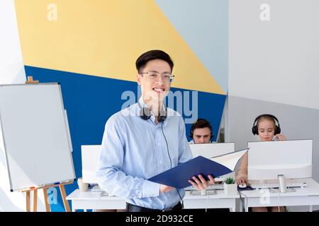 Happy Asian male helpdesk operator in headset holding documents at modern office Stock Photo