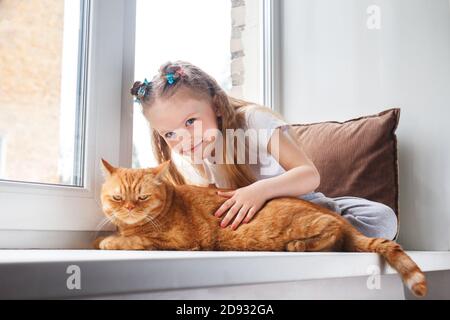 Cute child embraces with tenderness and love a red cat on window Stock Photo