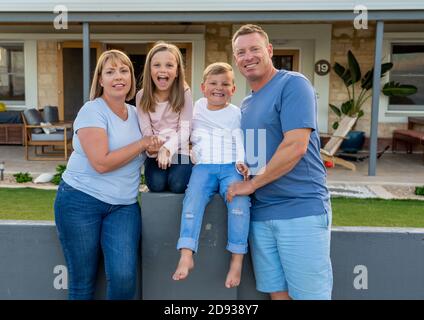 Outdoors portrait of a happy family of four smiling in front of new dream home or vacation rental house. Mom, dad, and children boy and girl, embracin Stock Photo