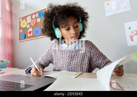 Smart african kid girl wears headphones learning online doing homework at home. Stock Photo