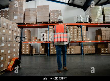 Back shot of male caucasian factory worker wearing overalls and hardhat standing by parcels in warehouse  Stock Photo