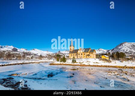 The wooden yellow Vågan Church on the beach on the Lofoten islands in Norway in winter with frozen river and mountains in morning light Stock Photo