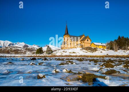 The wooden yellow Vågan Church on the beach on the Lofoten islands in Norway in winter with frozen river and mountains in morning light Stock Photo