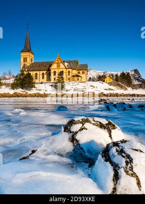 The wooden yellow Vågan Church on the beach on the Lofoten islands in Norway in winter with frozen river and mountains in morning light Stock Photo