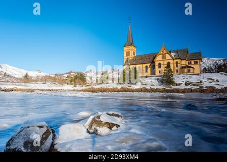 The wooden yellow Vågan Church on the beach on the Lofoten islands in Norway in winter with frozen river and mountains in morning light Stock Photo