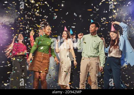 Group of young happy multiracial people wearing birthday hats and dancing, confetti falling in the air. Holiday, party and celebration concept Stock Photo