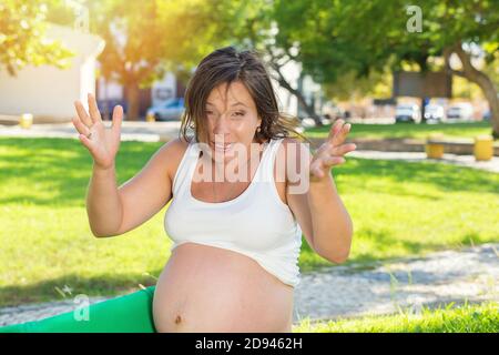Frustrating pregnant lady. Photo of emotional angry screaming pregnant woman hands in air in frustration sitting outside, outdoors in a park on a gree Stock Photo