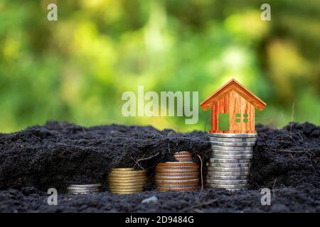Wooden house model on coin contains coins growing in the ground with money saving concept, green nature background. Stock Photo