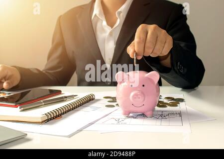 A business woman who is putting coins in a piggy bank, including a notebook on a wooden desk for work, ideas for saving money and managing accounts. Stock Photo