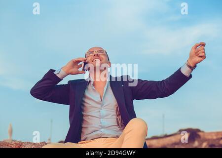 Businessman getting phenomenal news on the phone sitting on rocks fists hands in the air celebrating outdoors blue sky on background. Positive face ex Stock Photo