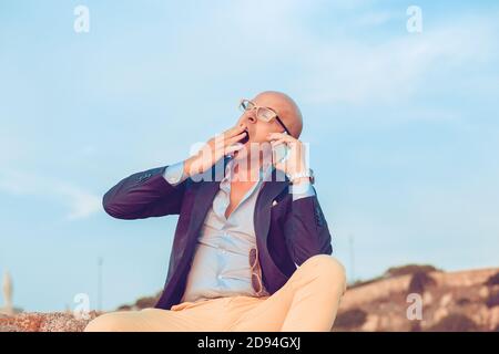 Tired yawning young bald business man banker dressed formally having coffee break outside city sitting outdoors blue sky on background. Funny face exp Stock Photo