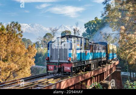 An approaching blue diesel train on railway tracks over a bridge in Himachal Pradesh, northern India with a view of the Himalayas Dhauladar Range Stock Photo